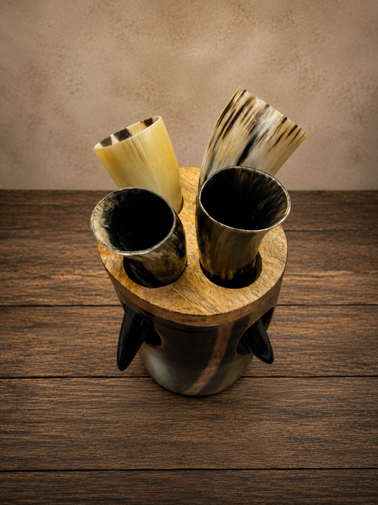 Four horn-shaped drinking cups in natural animal-hide tones, displayed in a round wooden holder with dark metal feet, set on a dark wood tabletop against a textured beige wall.